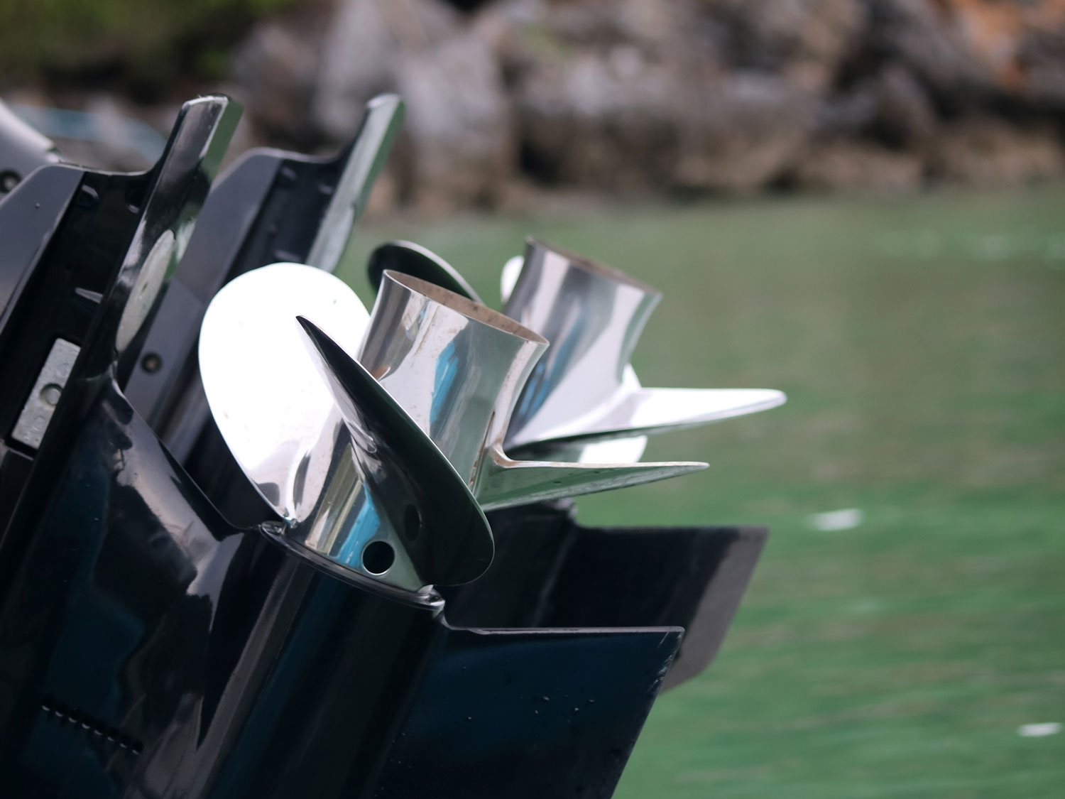 Two polished stainless steel outboard propellers on black engine lower units, with green water in the background.