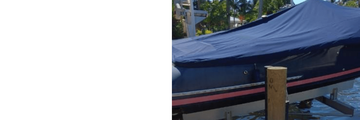 Navy blue boat under a fitted cover on a dock-side lift with a red bottom stripe, with palm trees and water in the background.