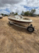 Small open motorboat with white and maroon hull resting on a bunk trailer on dry dirt ground.