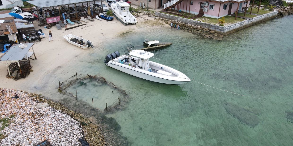 Aerial view of a white center-console sportfishing boat moored at a sandy shoreline beside a fish pen and beach bar, with a pink house behind the seawall.