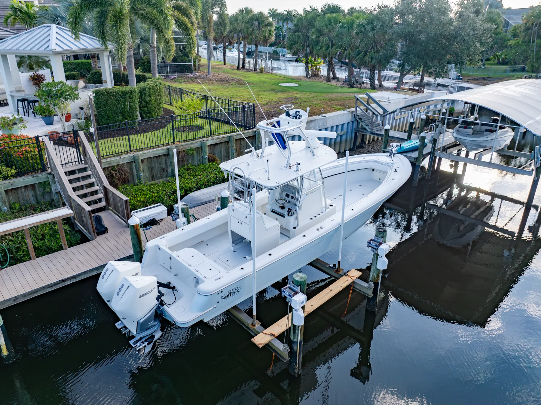 White Nords Boatworks center console with twin Mercury outboards on a boat lift at a residential canal dock with palm trees and a covered boat port beside it.