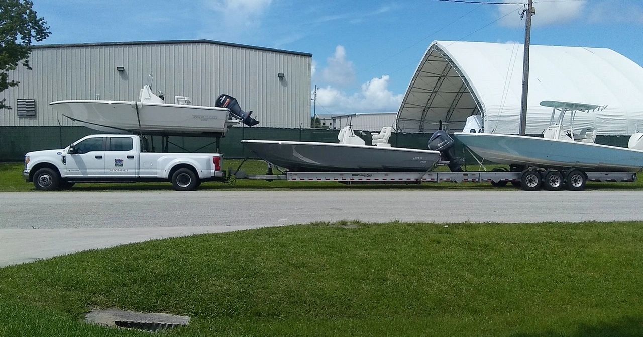 Three boats being hauled on a multi-boat aluminum trailer behind a white Ford dually pickup on a paved lot, with a white storage building and boat shelter in the background.