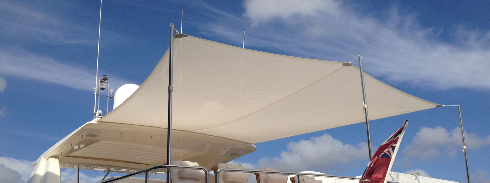 Cream fabric sun shade canopy over the flybridge seating of a motor yacht, with stainless steel support poles and a red ensign flag.
