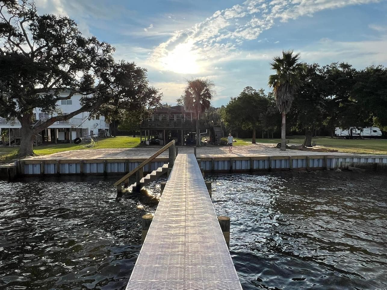 Newly built dock walkway extending over a river toward a waterfront yard with a timber-and-metal seawall at an unknown business, unknown town.