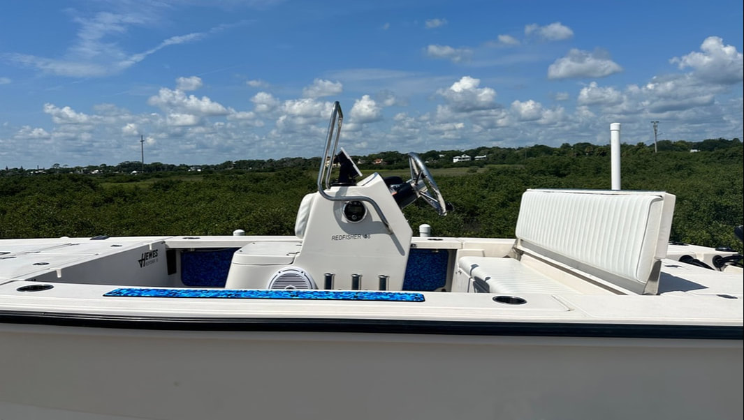 Bow and helm area of a white Redfisher flats boat with rod holders and a bench seat, with marsh and trees in the background.