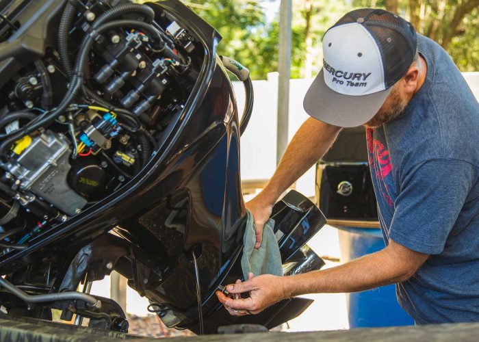 Technician cleaning a black outboard motor with a cloth while wearing a Mercury Pro Team cap, outdoors at a marine service location.