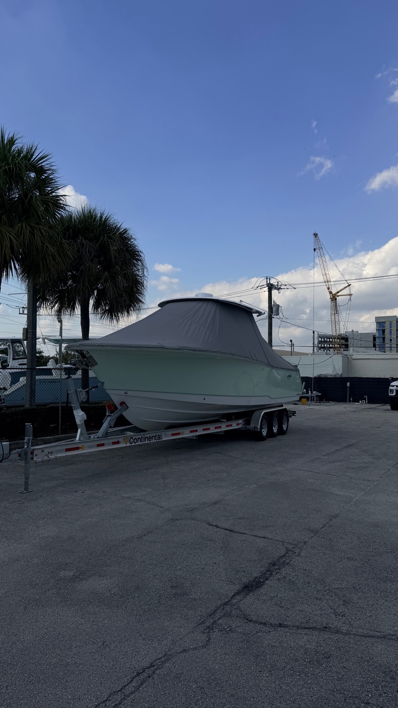 Sportfishing boat with a gray canvas T-top enclosure on a Continental trailer in a paved lot with palm trees and a construction crane nearby.