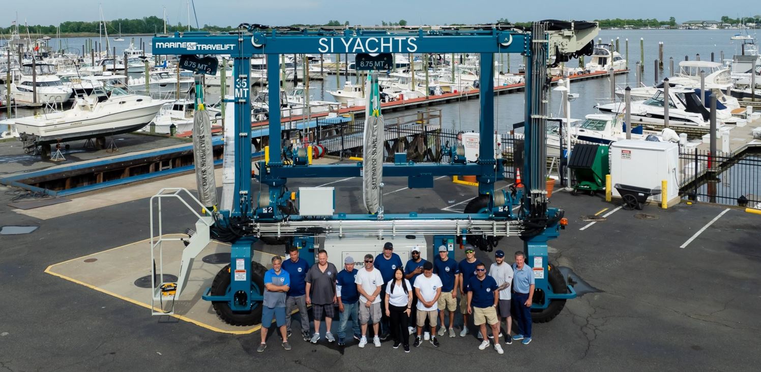 Crew members posing in front of a Marine Travelift boat hoist labeled SI Yachts at a marina with boats in the background.
