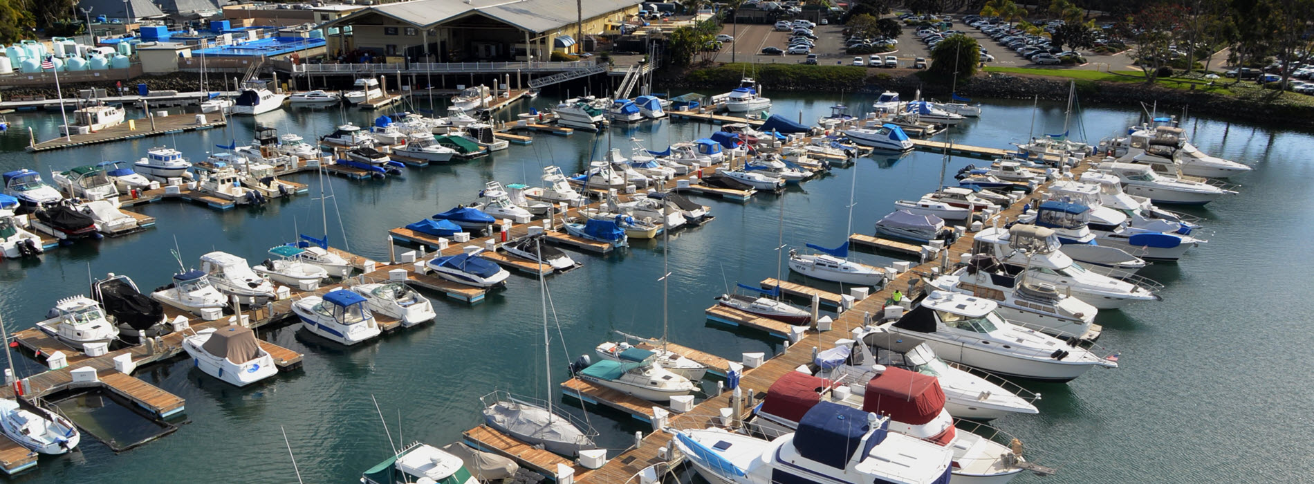 Aerial view of docked powerboats and sailboats at a harbor marina with a waterfront building and parking lot.
