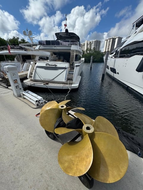 Brass propeller on a dock beside a white motor yacht in a canal, with high-rise buildings in the background.