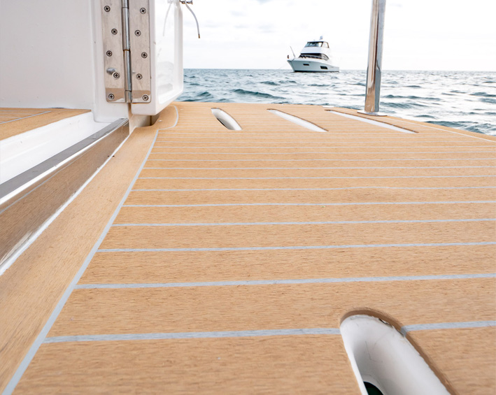 Synthetic teak-style deck planking on a boat stern with a white motor yacht on the water beyond.
