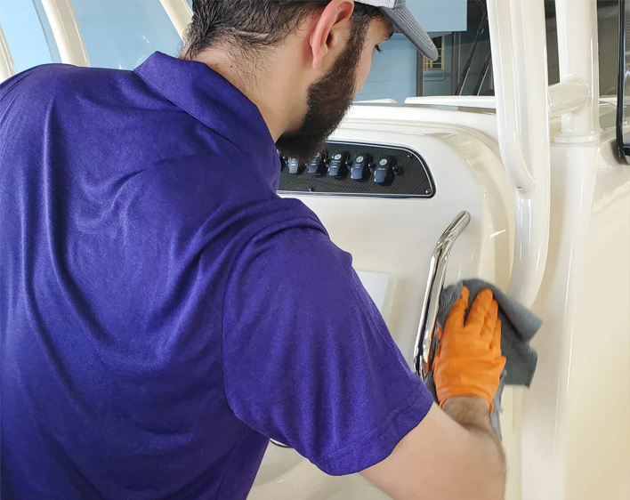 Technician in a purple polo and orange gloves wiping down the white helm console and chrome grab rail of a boat.