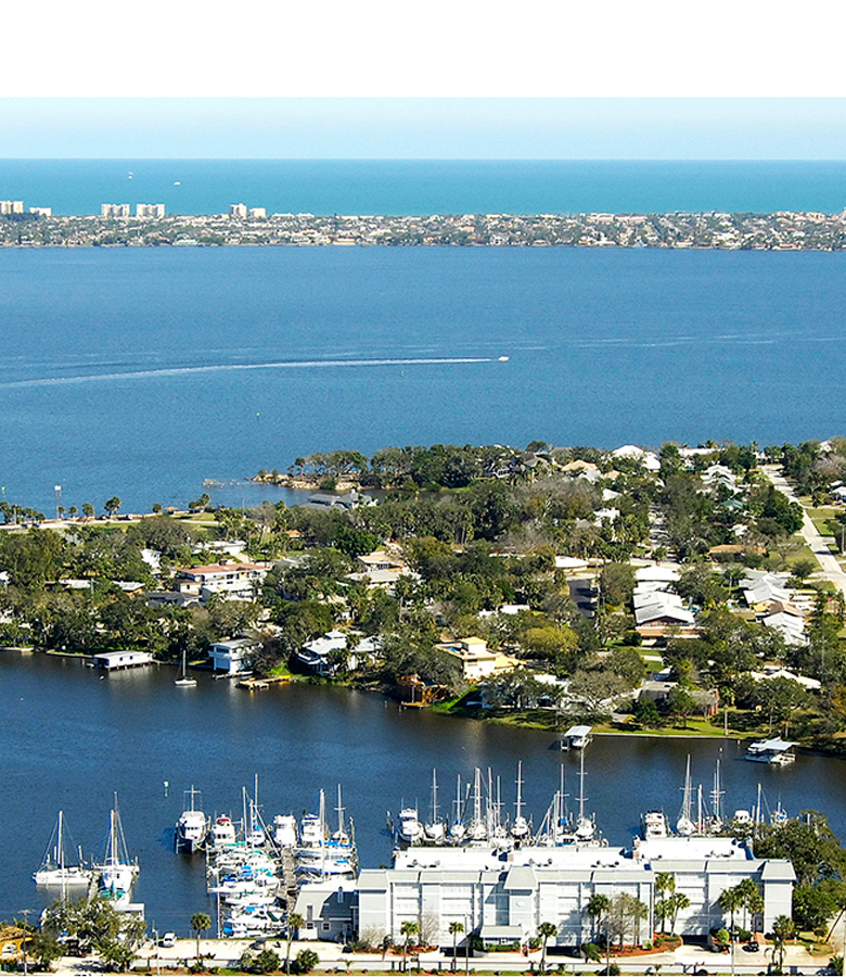 Aerial view of a marina with docked sailboats and powerboats beside a waterfront residential neighborhood, with a distant city skyline across the bay.