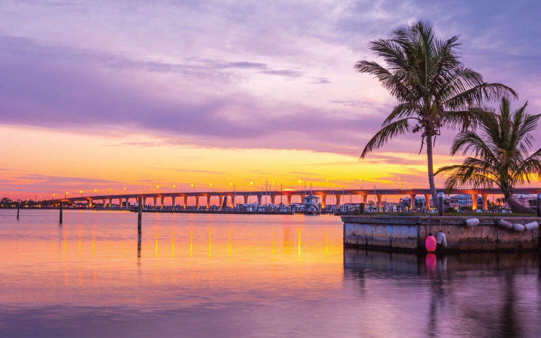Sunset view of a long causeway bridge over calm water with palm trees and a marina waterfront, location unidentified.