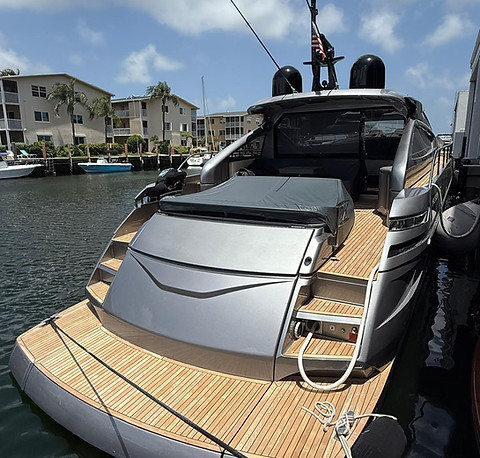 Stern of a gray motor yacht with a teak swim platform and cockpit seating, moored at a canal-side marina with condominiums in the background.