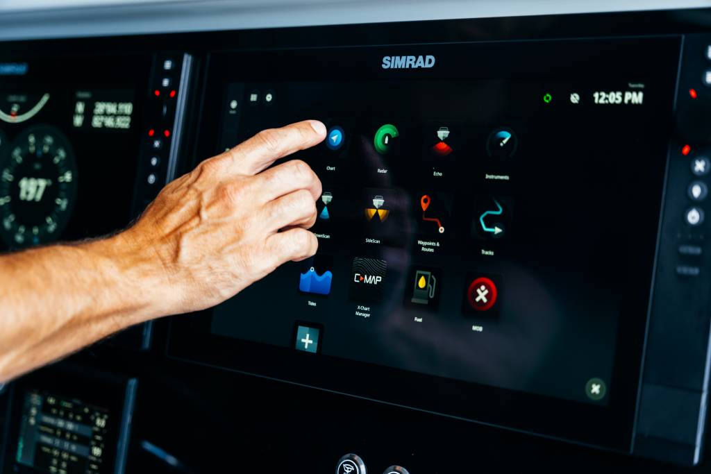 Hand operating a Simrad marine navigation touchscreen display with chart, radar, and sonar app icons at the helm of a boat.