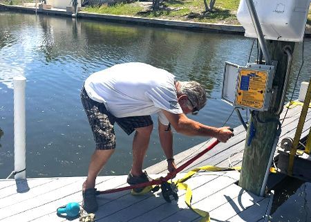 Man working with a red strap on a dock-side boat lift control box along a canal, with no business name or town visible.