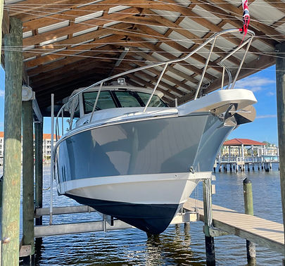 Motor yacht with a white hull and navy boot stripe resting on a covered boat lift at a marina, with a dock and waterfront buildings in the background.