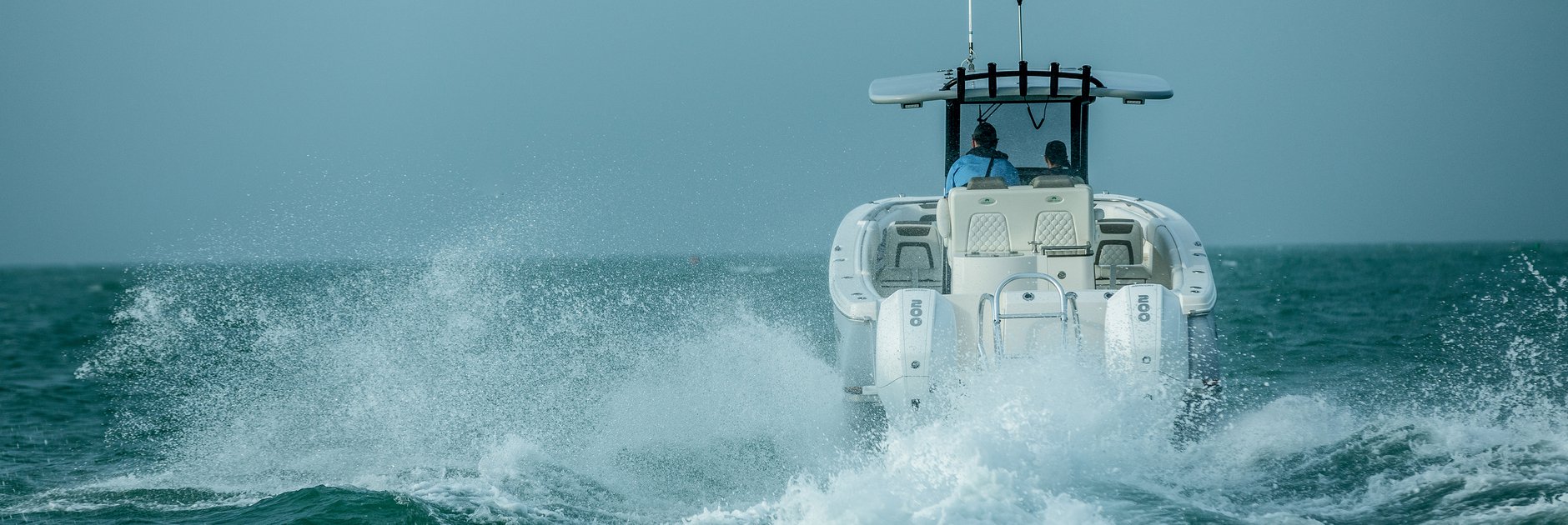 Center console boat with twin outboard motors kicking up a spray wake across open water, with two people aboard at the helm.