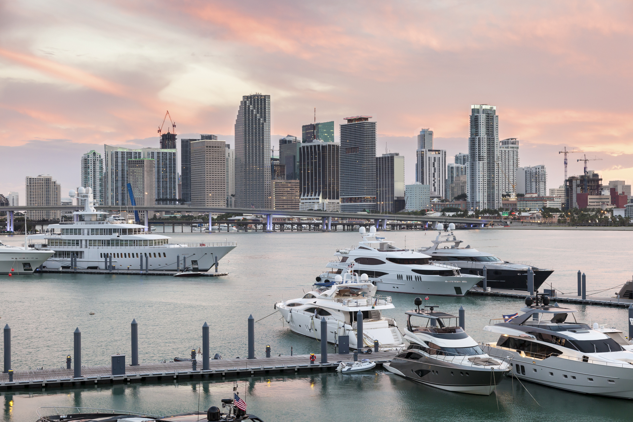 Luxury motor yachts docked at a marina with the Miami skyline behind them at sunset.