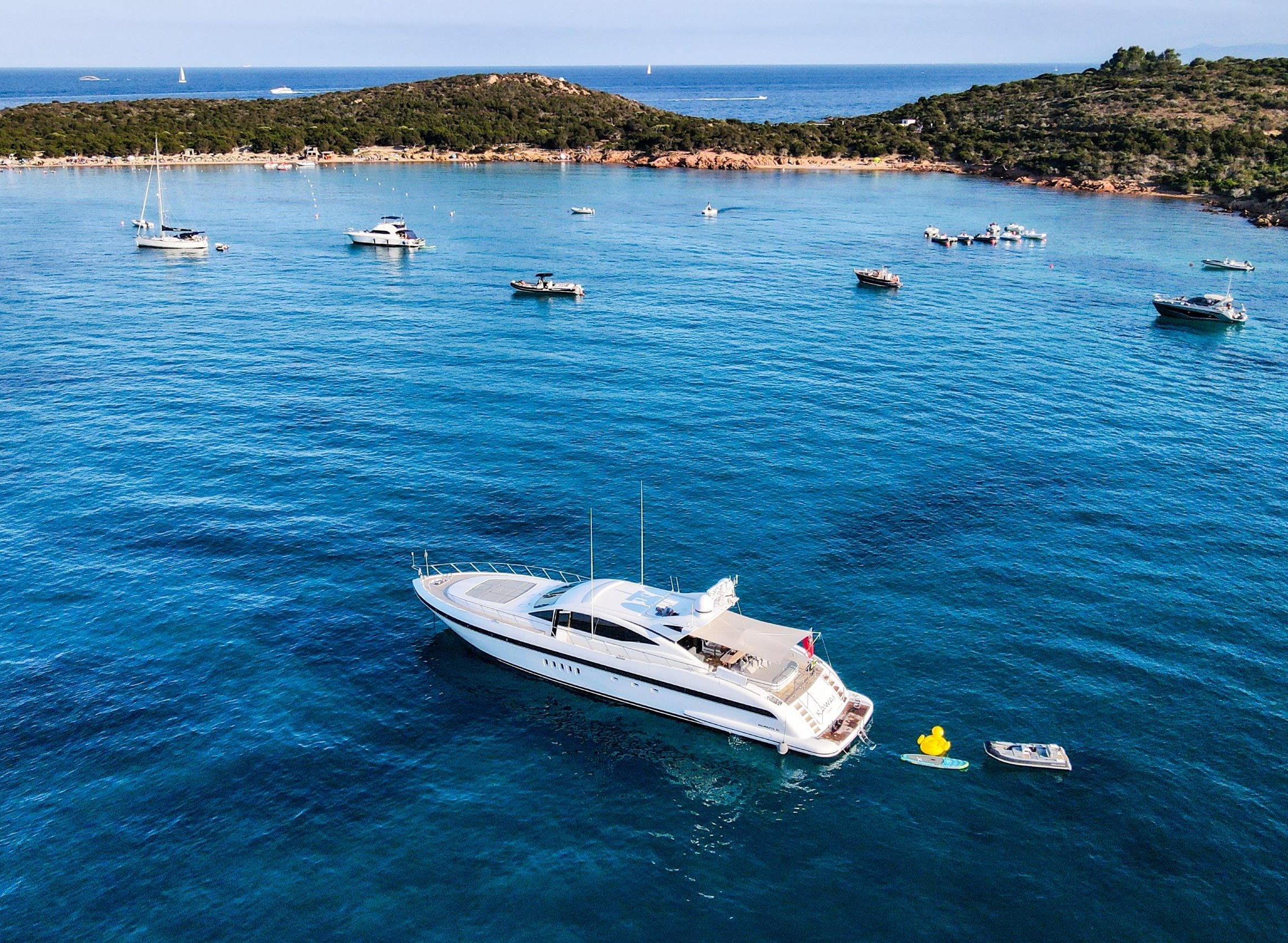 Aerial view of a white motor yacht anchored in a turquoise bay beside sailboats, tenders, and a wooded coastline.