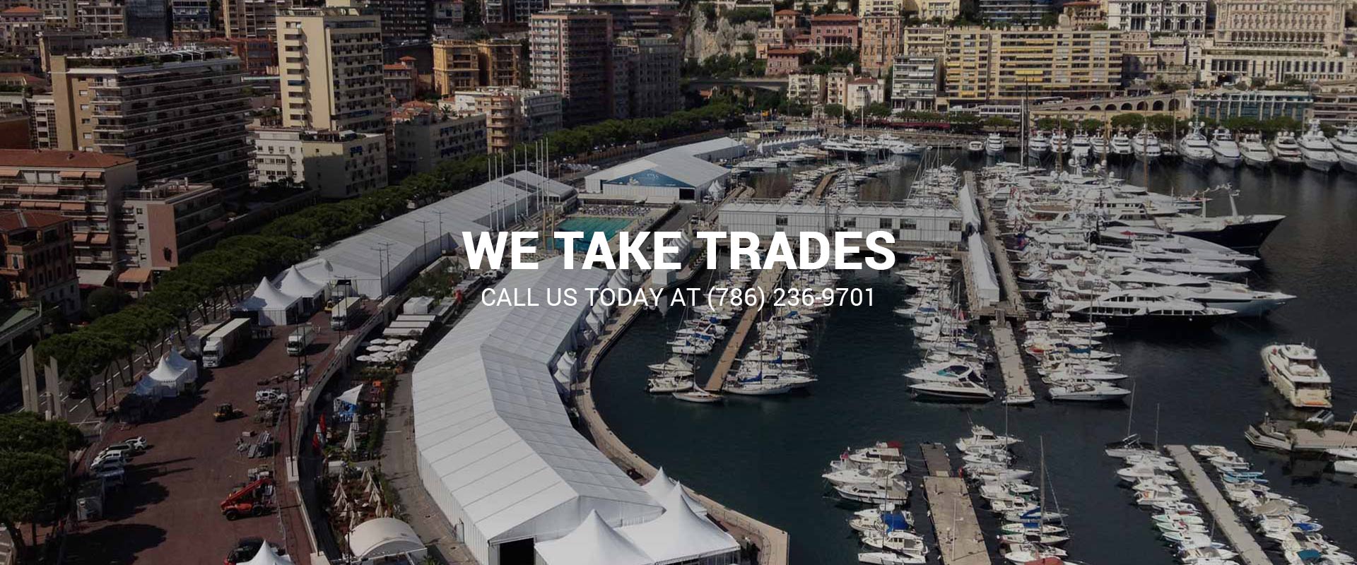 Aerial view of yachts and sailboats moored in a marina beside a large white event tent along the waterfront, with a hillside city behind, overlaid with a 'We Take Trades' banner.