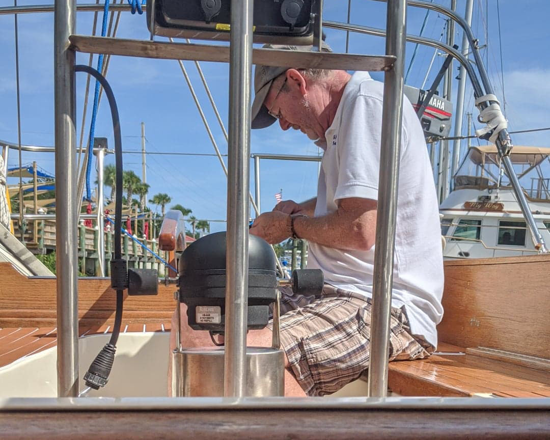 Boat owner working on a helm compass on the wooden deck of a sailboat at a marina dock.