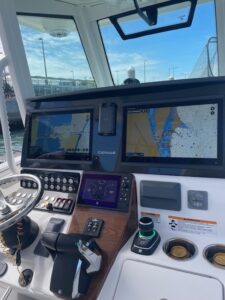 Boat helm with twin chartplotter screens, steering wheel, and engine controls at a marina.