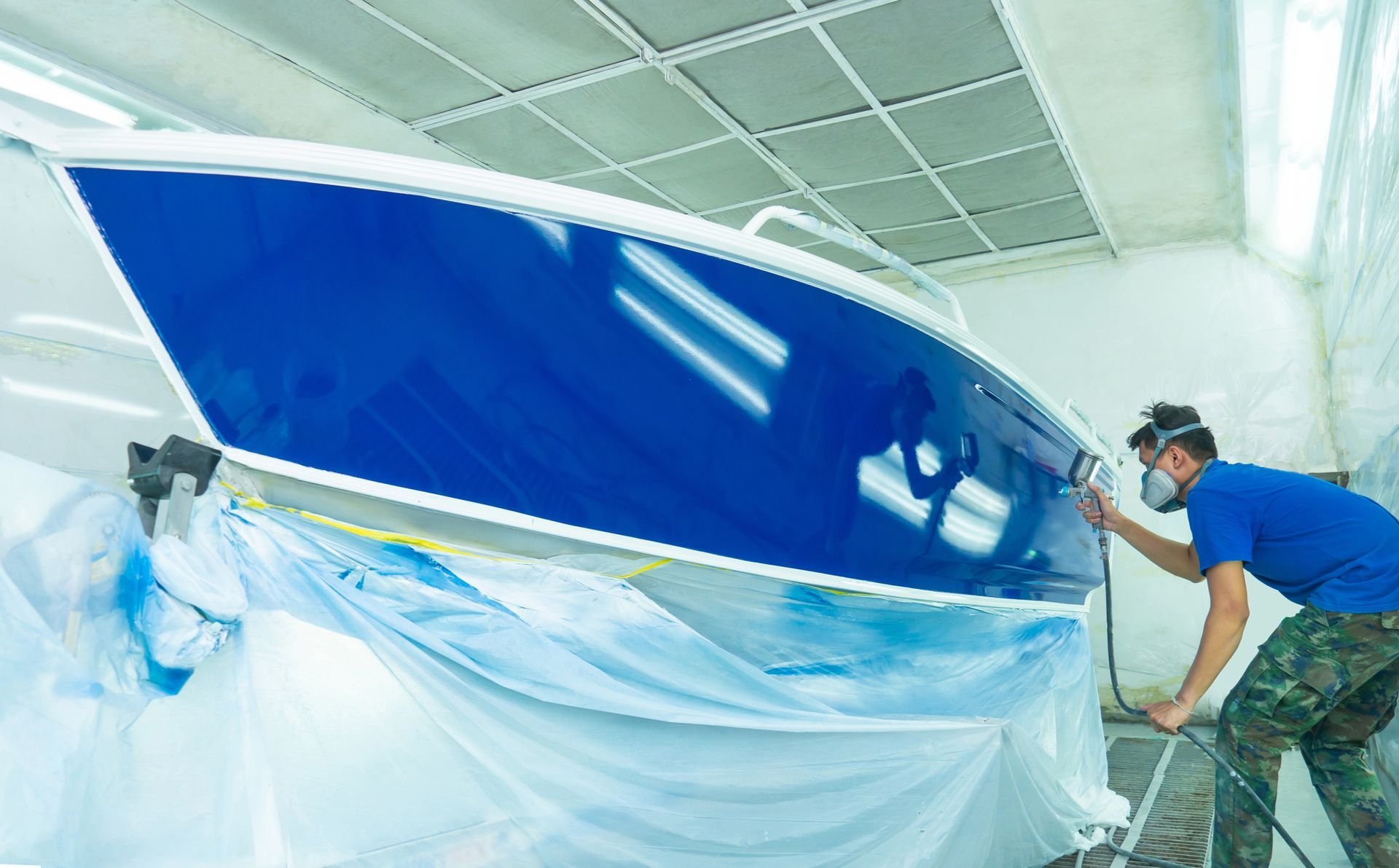 Technician in a respirator spray-painting a blue boat hull in a paint booth.