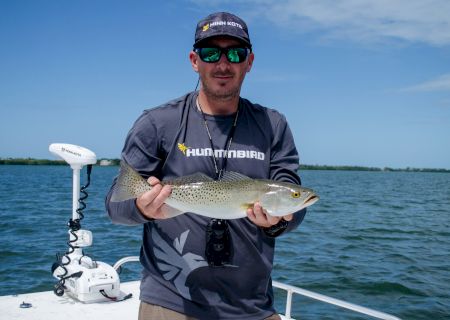 Angler in a Humminbird shirt holding a speckled sea trout on a boat with a bow-mounted trolling motor over calm bay water.