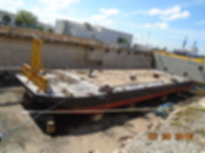 Flat-deck barge on a shipyard slipway with a yellow crane arm and industrial buildings behind it.