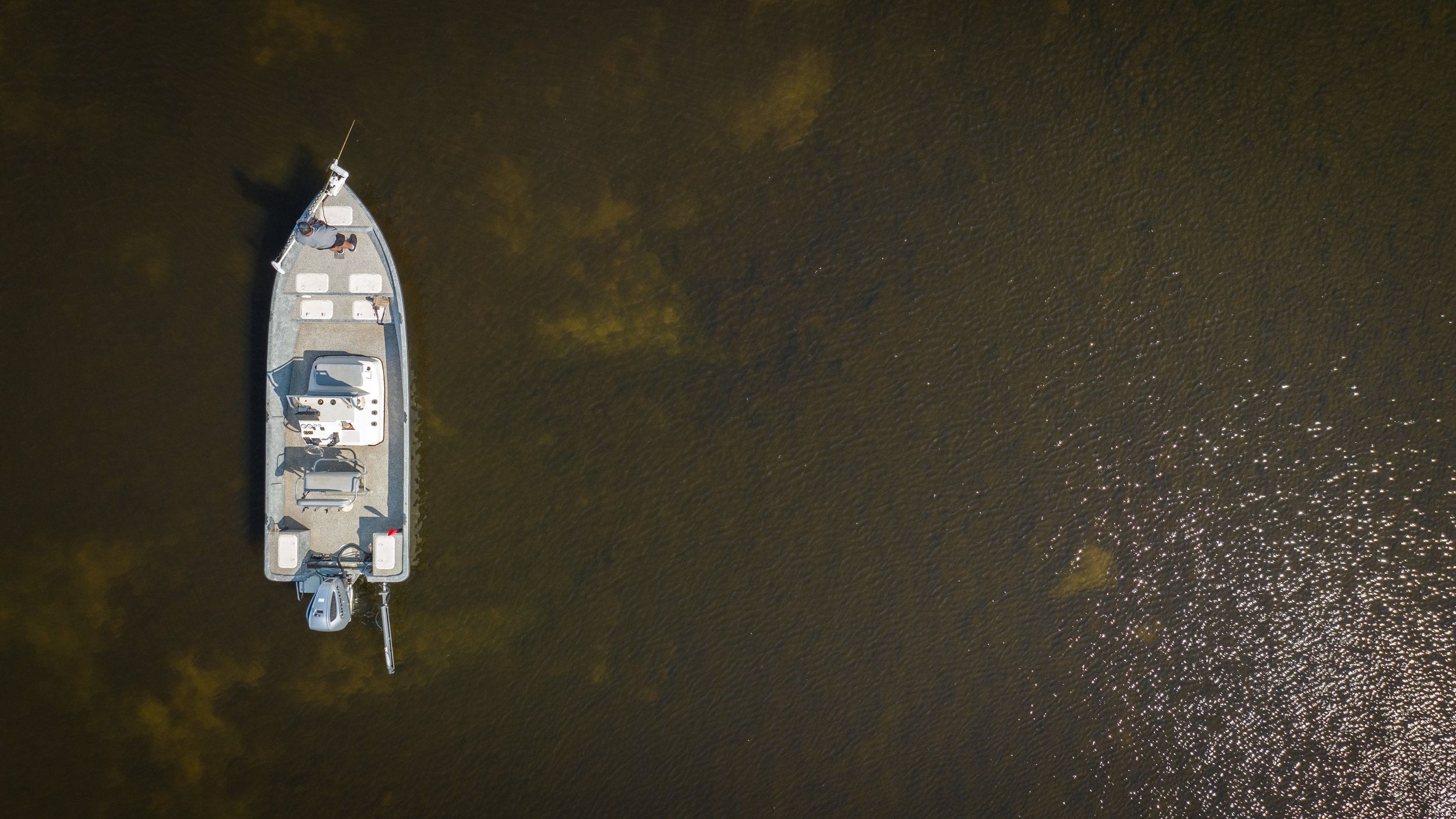 Aerial view of a small aluminum fishing boat with an outboard motor and an angler at the bow on calm water.