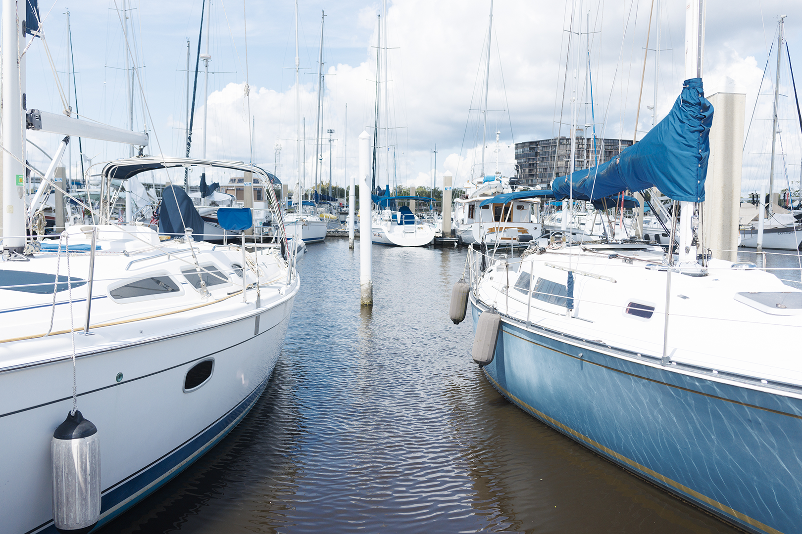Sailboats moored along a narrow marina channel with white pilings and fenders, under a partly cloudy sky.