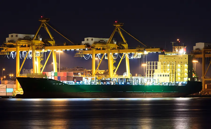 Container ship moored beside yellow gantry cranes and stacked containers at a port terminal at night.