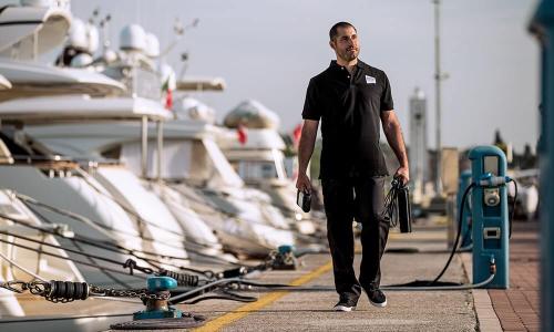 Crew member in a black polo walking along a marina pier past large white motor yachts and mooring cleats.