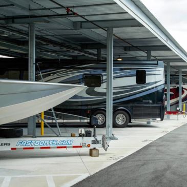 Covered outdoor storage bays holding a motorhome, a white boat hull, and a boat trailer marked fastboats.com, business and town unknown.