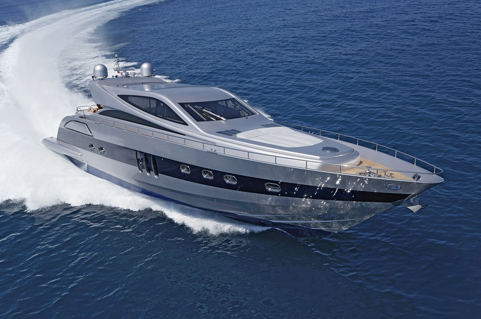 Silver motor yacht with a dark blue hull stripe under way at speed across open water, shown from an elevated angle.