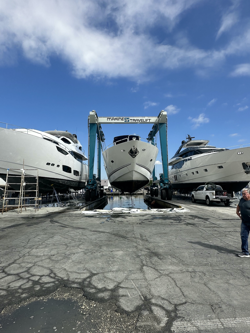Yacht being lifted by a Marine Travelift gantry between two motor yachts at a boatyard, town unknown.