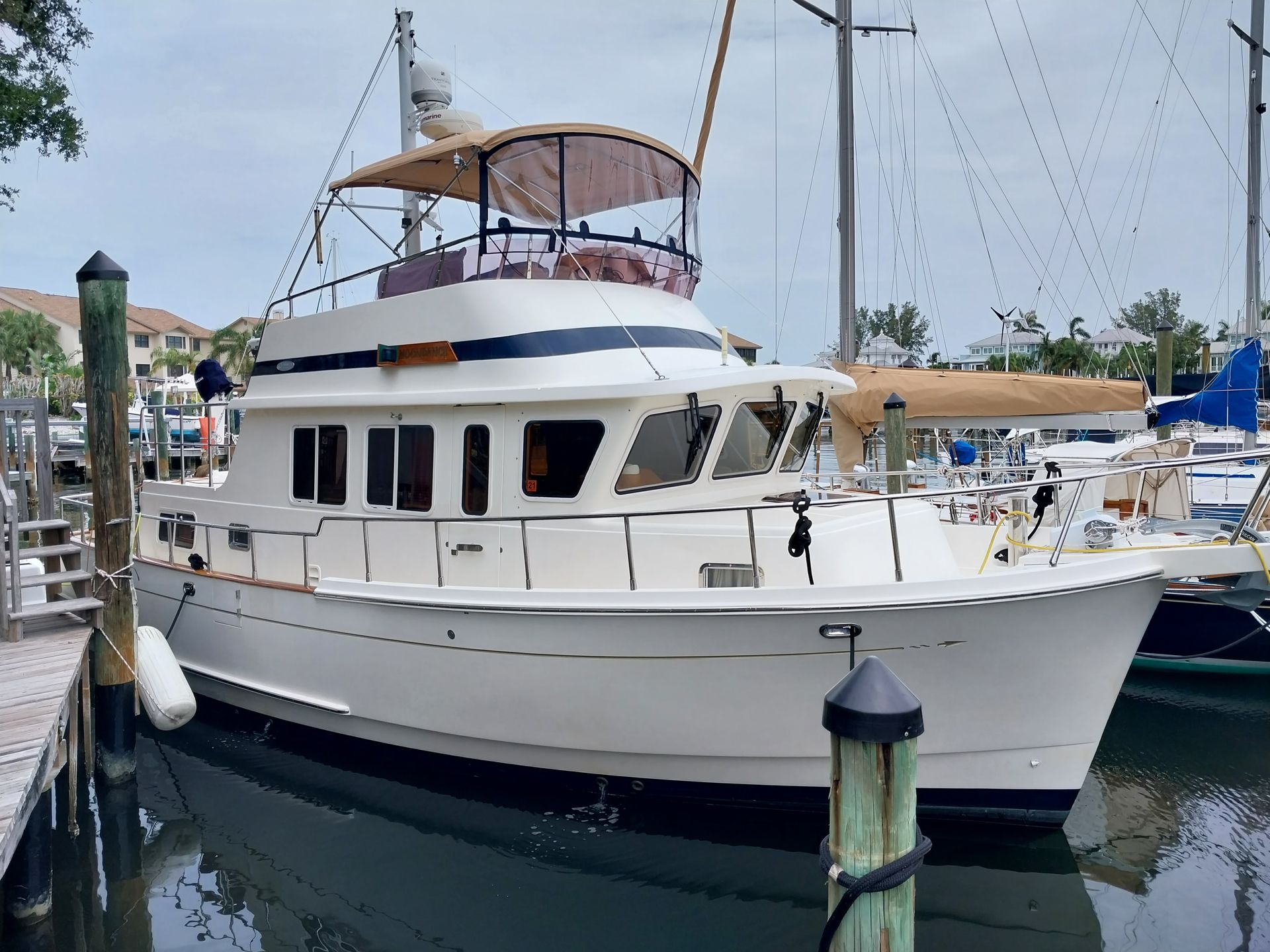 White motor yacht with a tan canvas bimini docked in a marina slip beside sailboats, with waterfront condos in the background.