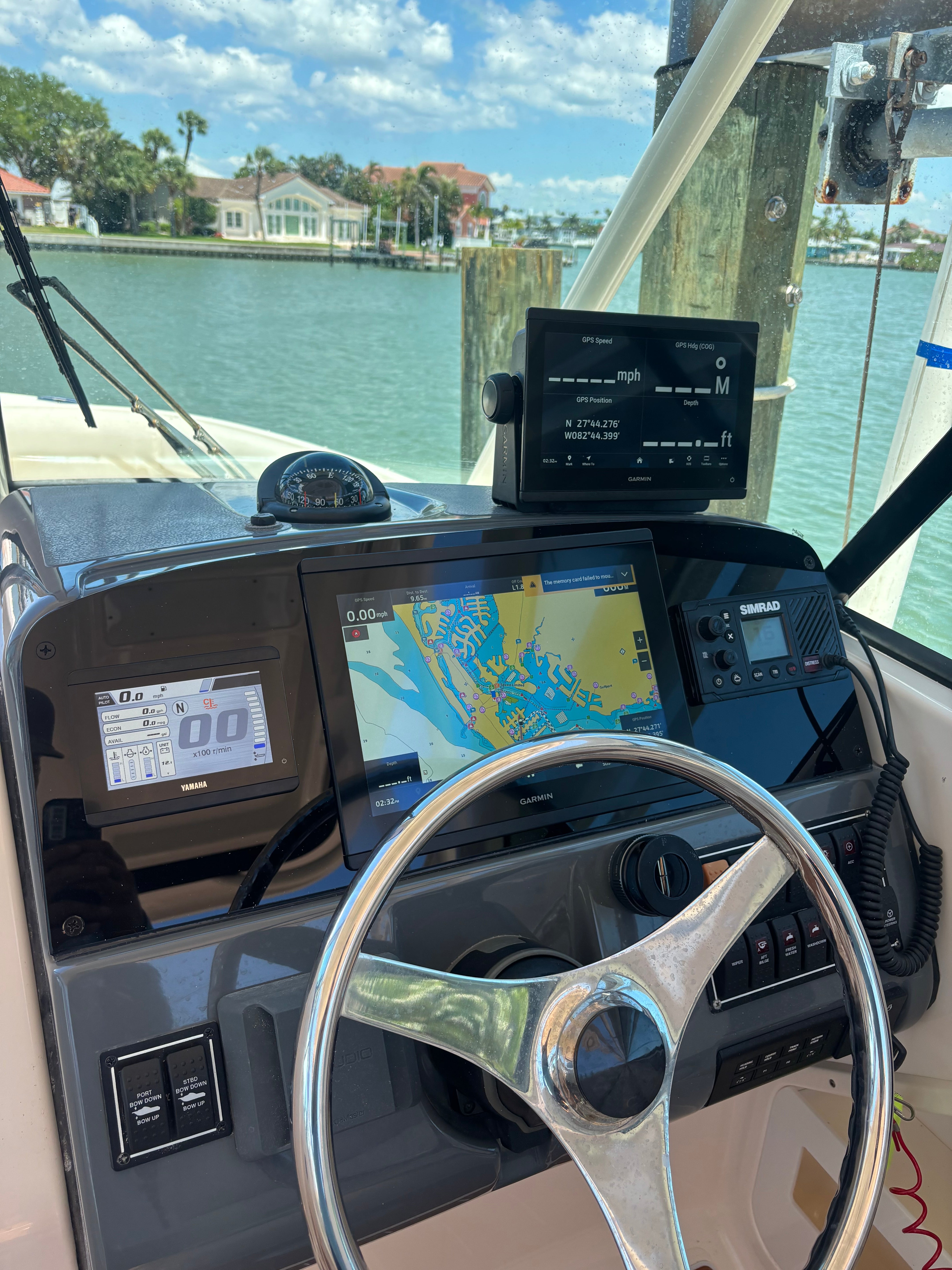 Boat helm with Garmin chartplotter, Simrad VHF radio and Yamaha engine display overlooking a residential canal, location not confirmed.