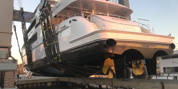 Large white motor yacht being lifted out of the water with straps at a boatyard.