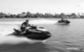 Two jet skis with riders speeding across open water near a tree-lined shoreline.