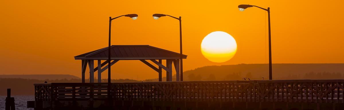 A wooden pier with a covered gazebo and street lamps silhouetted against a large orange sunset over the water.