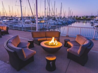 Outdoor fire pit lounge with curved sofas overlooking a marina full of docked sailboats at dusk.