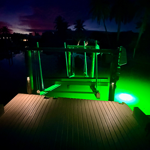 Boat lift and dock illuminated by green underwater lights at dusk on a canal waterfront.
