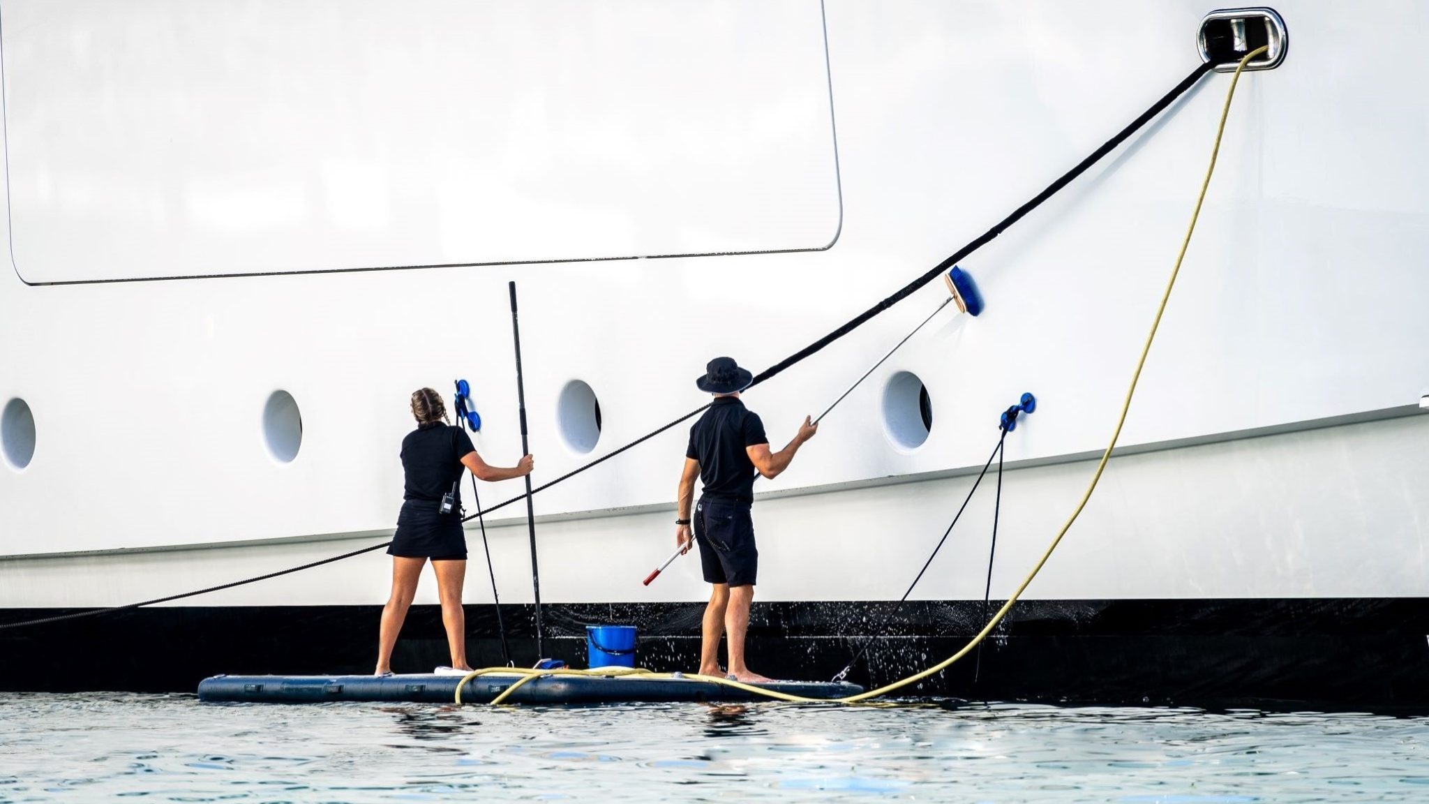 Two crew members hand-washing the white hull of a large yacht from a floating work platform on calm water.