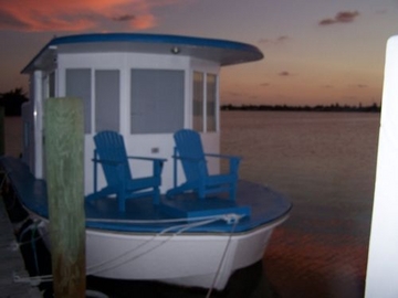 Small houseboat with blue Adirondack chairs on its deck tied at a dock at sunset, with no business name or town visible.