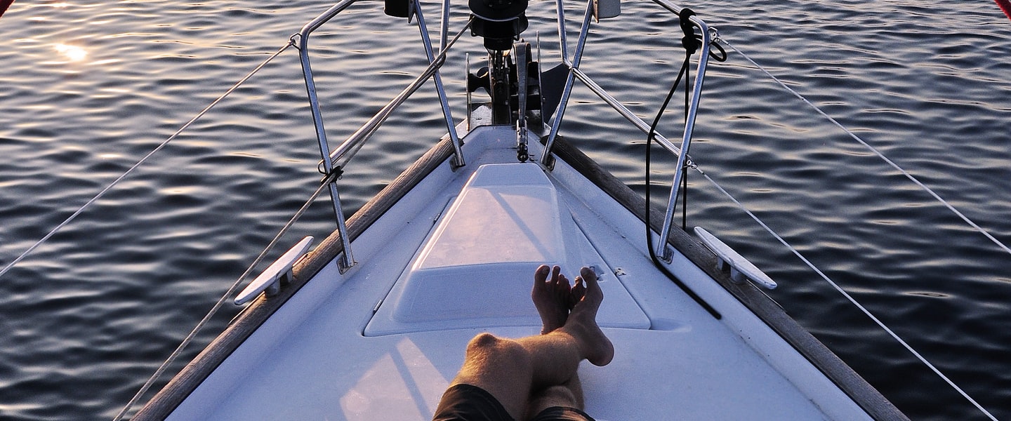 Bare feet resting on the foredeck of a sailboat at sunset, with stainless steel bow rails and calm water ahead.