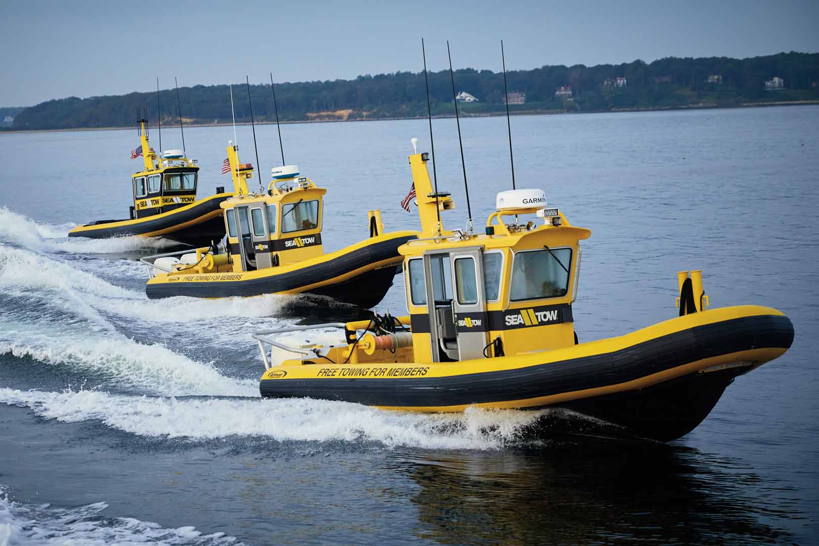 Three yellow Sea Tow rescue boats underway in formation on open water with a wooded shoreline behind them.