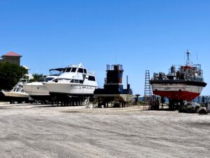 Motor yachts and a red-hulled workboat on stands at a boatyard hard-standing area, with no business name or town visible.
